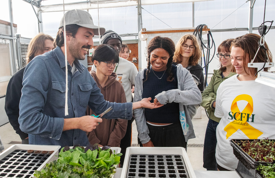 A farmer hands a plant clipping to a student as several classmates gather around in a greenhouse